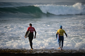 Two surfers wearing wetsuits and carrying their surfboards stand on the edge of the water, looking towards the powerful waves in the ocean. One is wearing a red top and the other a yellow top with a blue helmet.