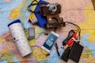 A collection of travel essentials sits on a detailed map. A vintage camera in a brown leather case rests near a smartphone displaying Cyrillic text. Next to it, a large white travel tumbler marked 'Journal' stands beside several electronic gadgets, including a portable music player, an external hard drive, and a set of coiled cables. A pack of color film and a blue notebook complete the setup.