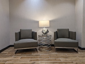 Two modern, dark gray upholstered armchairs are positioned in a symmetrical layout on either side of a metal and glass cylindrical side table with a decorative lamp. The setting has a minimalist aesthetic, featuring wood-look tile flooring and neutral-colored walls.