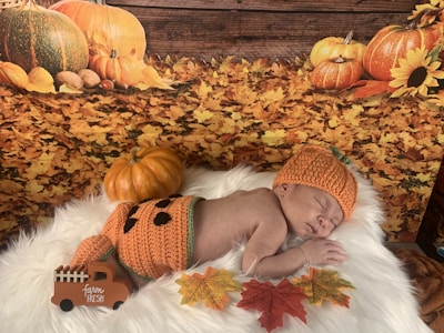 A baby is peacefully sleeping on a soft white fur surface, dressed in an orange knitted outfit that resembles a pumpkin, complete with a pumpkin hat. Surrounding the baby are autumn-themed decorations, including a small pumpkin, colorful fall leaves, and a wooden board with a 'farm fresh' toy truck. A backdrop of fallen leaves and pumpkins enhances the autumn atmosphere.