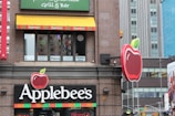 A building facade of an Applebee's restaurant with distinct branding, including large red apple logos and colorful striped decorations. The windows display promotional signs for pasta and happy hour. The surrounding architecture consists of modern urban buildings.