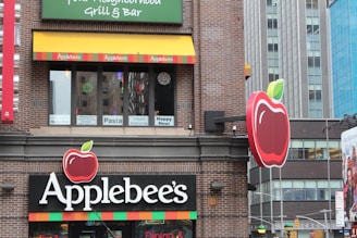 A building facade of an Applebee's restaurant with distinct branding, including large red apple logos and colorful striped decorations. The windows display promotional signs for pasta and happy hour. The surrounding architecture consists of modern urban buildings.
