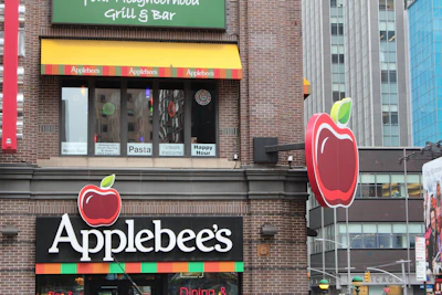A building facade of an Applebee's restaurant with distinct branding, including large red apple logos and colorful striped decorations. The windows display promotional signs for pasta and happy hour. The surrounding architecture consists of modern urban buildings.