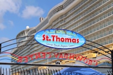 A large cruise ship is docked, with a sign in the foreground reading 'St. Thomas - Come Back Again Soon!' The ship features multiple balconies and appears luxurious. The sky is clear with a few clouds, suggesting a pleasant day.