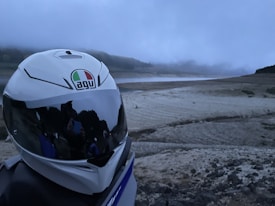 A motorcycle helmet with a reflective visor displaying a logo is positioned in the foreground on an off-road setting. In the background, a misty, overcast sky blankets a barren, rocky landscape.