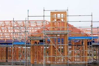 A partially constructed building framework is surrounded by metal scaffolding. The timber structure includes beams and trusses, indicating ongoing construction work. The framework is set against a clear sky, highlighting an early stage in the building process.