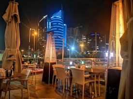 An outdoor dining area with wicker chairs and tables is set up on a terrace with tall outdoor heaters. In the background, there's a view of a cityscape at night, featuring modern skyscrapers illuminated with vibrant blue lighting and a busy street with traffic and streetlights.