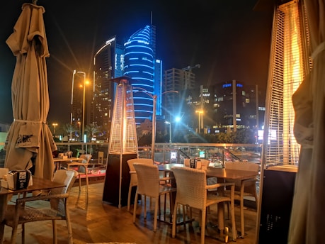 An outdoor dining area with wicker chairs and tables is set up on a terrace with tall outdoor heaters. In the background, there's a view of a cityscape at night, featuring modern skyscrapers illuminated with vibrant blue lighting and a busy street with traffic and streetlights.