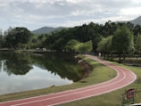 A serene landscape featuring a winding red running track alongside a calm lake surrounded by lush green trees. The scene includes reflections of the trees on the water, under a partly cloudy sky with distant hills in the backdrop.