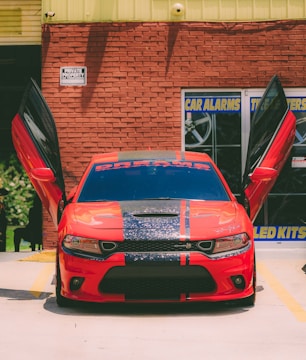 A vivid red sports car parked in front of a brick wall with both its gullwing doors open. The car has black racing stripes down the middle of its hood and roof, and a decal on the windshield that appears to read 'DREAMS.' The background includes a building with signage related to car alarms and kits.
