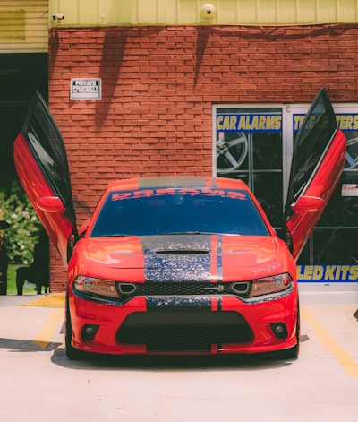 A vivid red sports car parked in front of a brick wall with both its gullwing doors open. The car has black racing stripes down the middle of its hood and roof, and a decal on the windshield that appears to read 'DREAMS.' The background includes a building with signage related to car alarms and kits.