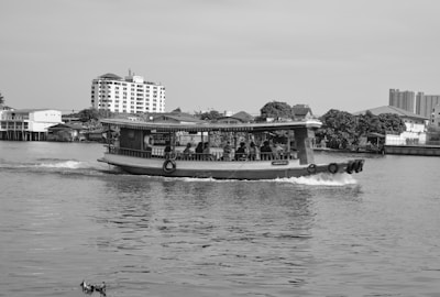 A traditional wooden boat is cruising on a calm river, with several passengers on board. In the background, there are residential buildings and lush green trees lining the riverbank under a clear sky. The scene is peaceful and captures a blend of urban and natural elements.