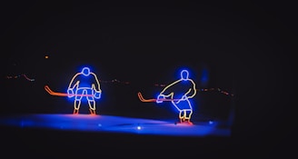 Two neon outlines of hockey players are illuminated against a dark background. Each figure is outlined in blue and yellow neon lights, with red sticks in their hands. The scene appears to be an artistic representation of a hockey game.