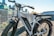 An electric bicycle with wide tires is positioned on a gravel pathway near a metal railing. The bike appears sturdy and suitable for off-road adventures. In the background, a modern cityscape is visible with several tall buildings under a clear blue sky.
