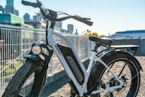 An electric bicycle with wide tires is positioned on a gravel pathway near a metal railing. The bike appears sturdy and suitable for off-road adventures. In the background, a modern cityscape is visible with several tall buildings under a clear blue sky.