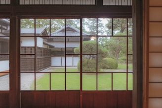 A serene view from inside a traditional Japanese building with wooden frames and panels, looking out through large windows onto a garden with manicured bushes, trees, and a stone path leading to another traditional structure. The architecture features dark wood and white walls, typical of Japanese design.