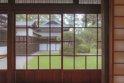 A serene view from inside a traditional Japanese building with wooden frames and panels, looking out through large windows onto a garden with manicured bushes, trees, and a stone path leading to another traditional structure. The architecture features dark wood and white walls, typical of Japanese design.