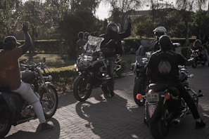 A group of motorcyclists gather on a sunlit street, with one person in a helmet raising their hand. The scene is set against a backdrop of trees and greenery, creating a casual, leisurely atmosphere.