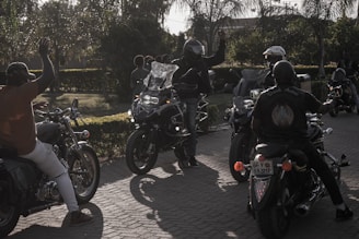 A group of motorcyclists gather on a sunlit street, with one person in a helmet raising their hand. The scene is set against a backdrop of trees and greenery, creating a casual, leisurely atmosphere.
