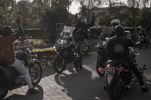 A group of motorcyclists gather on a sunlit street, with one person in a helmet raising their hand. The scene is set against a backdrop of trees and greenery, creating a casual, leisurely atmosphere.