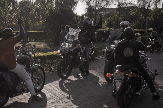 A group of motorcyclists gather on a sunlit street, with one person in a helmet raising their hand. The scene is set against a backdrop of trees and greenery, creating a casual, leisurely atmosphere.