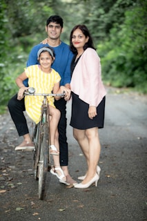 A family of three stands on a tree-lined road, with the father in a blue shirt sitting on a bicycle and the daughter in a yellow striped dress sitting on the bicycle handle. The mother is standing next to them wearing a pink blazer over a black dress. The backdrop is a lush, green natural setting.