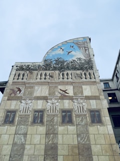 A painted building facade with architectural elements such as columns and windows, featuring an upper section depicting birds flying in a sky backdrop among leaves and decorative curtains.