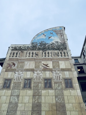 A painted building facade with architectural elements such as columns and windows, featuring an upper section depicting birds flying in a sky backdrop among leaves and decorative curtains.