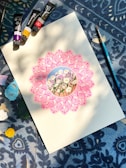 A detailed drawing of daisies is surrounded by an intricate pink mandala on a piece of paper. Paint tubes and a brush are arranged on a patterned blue fabric background, with patches of various colors around.