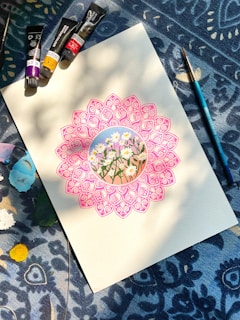 A detailed drawing of daisies is surrounded by an intricate pink mandala on a piece of paper. Paint tubes and a brush are arranged on a patterned blue fabric background, with patches of various colors around.