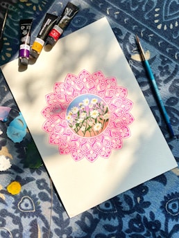 A detailed drawing of daisies is surrounded by an intricate pink mandala on a piece of paper. Paint tubes and a brush are arranged on a patterned blue fabric background, with patches of various colors around.