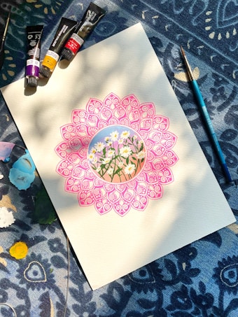 A detailed drawing of daisies is surrounded by an intricate pink mandala on a piece of paper. Paint tubes and a brush are arranged on a patterned blue fabric background, with patches of various colors around.