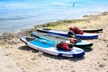 Several paddleboards are lined up on a sandy beach, each equipped with red life jackets and other gear. The boards are positioned near the water's edge with clear blue water in the background. A distant swimmer can be seen enjoying the sea. The scene captures a vibrant, sunny day ideal for water activities.