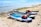 Several paddleboards are lined up on a sandy beach, each equipped with red life jackets and other gear. The boards are positioned near the water's edge with clear blue water in the background. A distant swimmer can be seen enjoying the sea. The scene captures a vibrant, sunny day ideal for water activities.