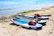 Several paddleboards are lined up on a sandy beach, each equipped with red life jackets and other gear. The boards are positioned near the water's edge with clear blue water in the background. A distant swimmer can be seen enjoying the sea. The scene captures a vibrant, sunny day ideal for water activities.
