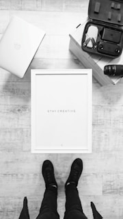 A top-down view of a minimalistic workspace setup featuring a closed laptop with an apple logo, an open drone case with a drone inside, a camera lens, and a framed piece of art with the words 'Stay Creative' at the center. A pair of feet in black shoes can be seen at the bottom of the image, standing on a light wooden floor.