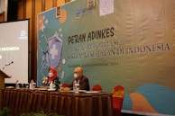 Two individuals are sitting at a conference table with microphones, one wearing a red headscarf. A large banner in the background displays text related to a health reform event in Indonesia. Several bottles and glasses are on the table, and a projection screen is visible to the left.