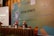 Two individuals are sitting at a conference table with microphones, one wearing a red headscarf. A large banner in the background displays text related to a health reform event in Indonesia. Several bottles and glasses are on the table, and a projection screen is visible to the left.