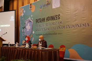 Two individuals are sitting at a conference table with microphones, one wearing a red headscarf. A large banner in the background displays text related to a health reform event in Indonesia. Several bottles and glasses are on the table, and a projection screen is visible to the left.
