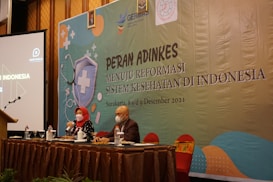 Two individuals are sitting at a conference table with microphones, one wearing a red headscarf. A large banner in the background displays text related to a health reform event in Indonesia. Several bottles and glasses are on the table, and a projection screen is visible to the left.