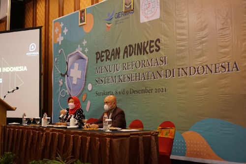 Two individuals are sitting at a conference table with microphones, one wearing a red headscarf. A large banner in the background displays text related to a health reform event in Indonesia. Several bottles and glasses are on the table, and a projection screen is visible to the left.