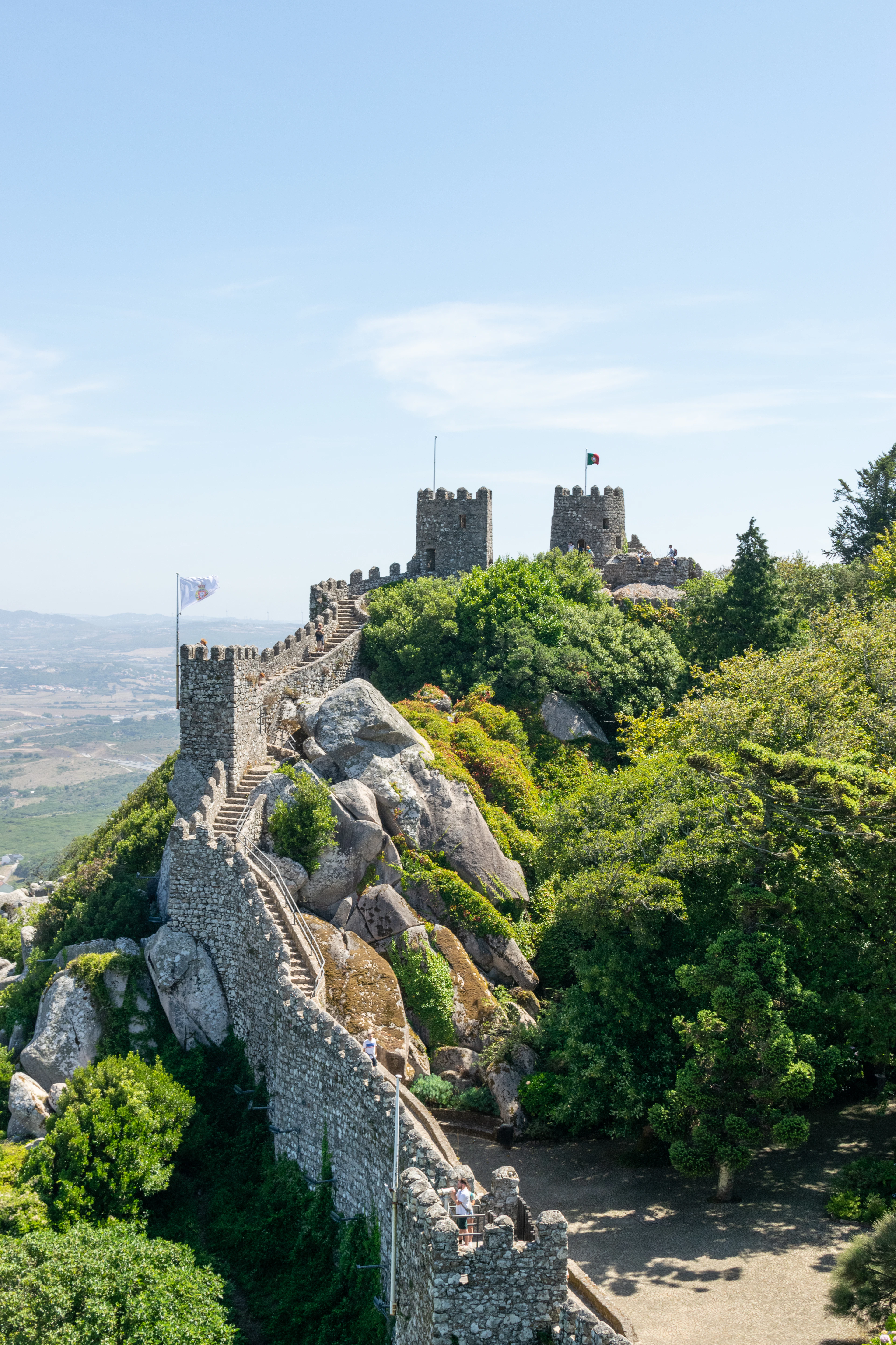 The battlemented stone walls of the Castle of the Moors in Sintra climbing over huge granite boulders to its two towers, the Portuguese flag flying above and the plain stretching away below