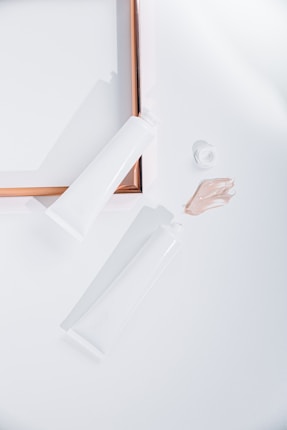 Two white tubes, one open with a small amount of beige cream or lotion next to it, are placed on a white surface. A rose gold picture frame is partially visible, creating a clean and minimalist arrangement.