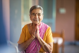 An elderly woman with gray hair is smiling warmly and standing with her hands together in a gesture of greeting or respect. She is wearing a colorful saree, combining shades of yellow and pink checkered patterns. The setting appears to be indoors with soft, natural lighting and blurred background.