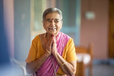 An elderly woman with gray hair is smiling warmly and standing with her hands together in a gesture of greeting or respect. She is wearing a colorful saree, combining shades of yellow and pink checkered patterns. The setting appears to be indoors with soft, natural lighting and blurred background.