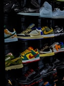 A display shelf features a collection of various sneakers, showcasing different brands and styles. The shoes are neatly arranged, with noticeable variations in color and design, including high-tops and low-tops. The lighting highlights the vibrant colors and logos on the sneakers.