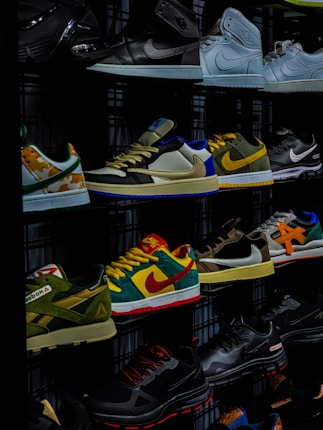 A display shelf features a collection of various sneakers, showcasing different brands and styles. The shoes are neatly arranged, with noticeable variations in color and design, including high-tops and low-tops. The lighting highlights the vibrant colors and logos on the sneakers.