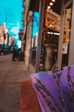 A bouquet of lavender flowers wrapped in purple paper is in the foreground, positioned on a street with a brightly lit storefront on the right side. The background shows blurred buildings and a bright blue sky, with hints of pedestrians or objects further along the sidewalk.