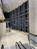 A modern indoor space features tall vertical windows with black framing, allowing views of the outside. The ceiling is high with a pattern of recessed lighting. The floor is tiled in beige and is connected to an escalator. There's a bench and potted plants present, creating a sleek, clean look.