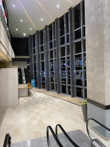 A modern indoor space features tall vertical windows with black framing, allowing views of the outside. The ceiling is high with a pattern of recessed lighting. The floor is tiled in beige and is connected to an escalator. There's a bench and potted plants present, creating a sleek, clean look.